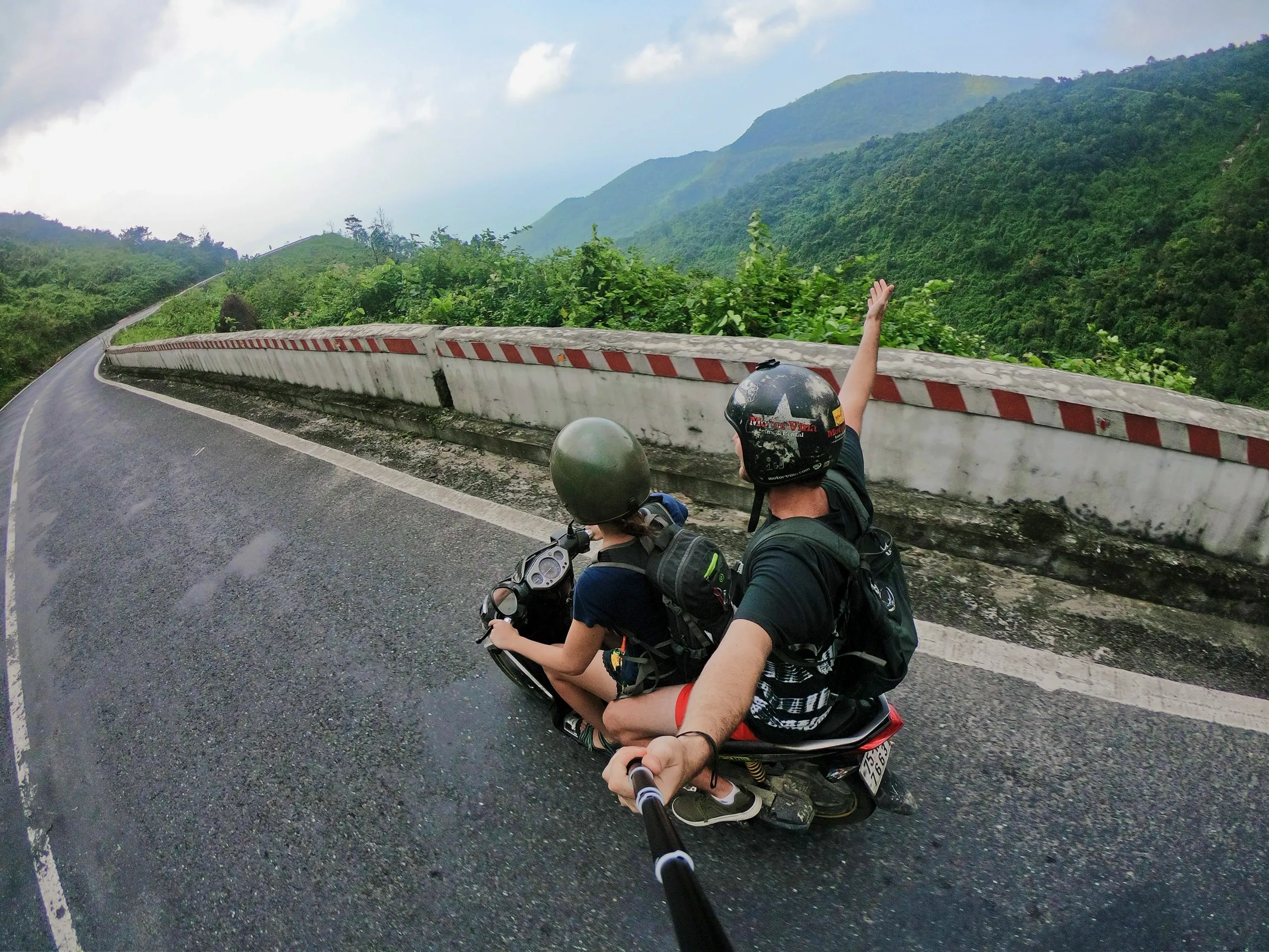 Two people on a motorcycle riding along a winding mountain road. One raises an arm excitedly. Lush green hills and cloudy sky in the background.