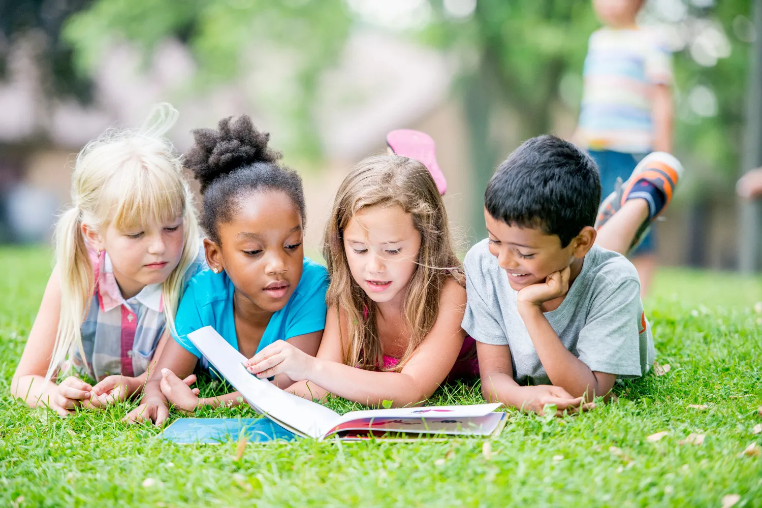 Four children lying on grass, reading a book together in a park. They’re focused and engaged. Background shows trees and blurred figures.
