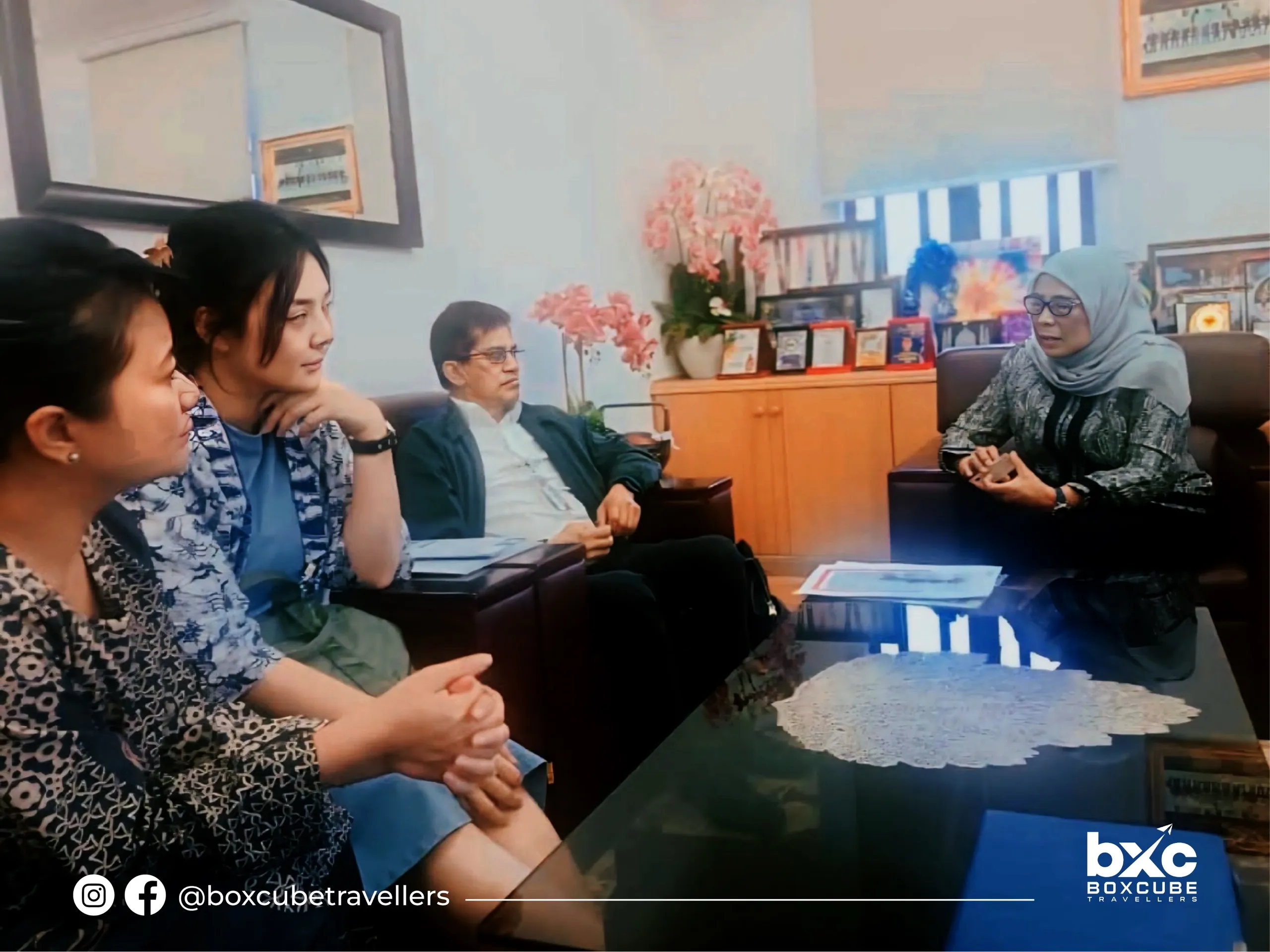 Four people are in a meeting in an office with a glass table, floral arrangements, and framed awards. They’re engaged in conversation.