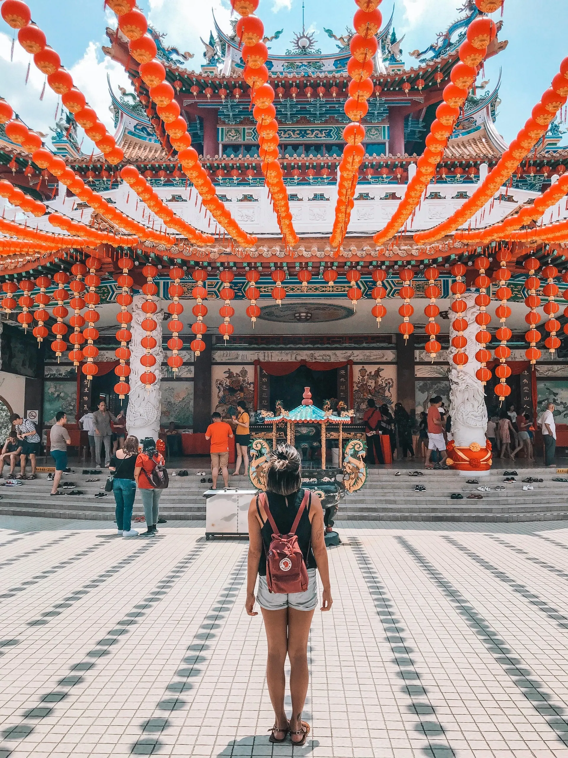 Thean Hou Temple in Malaysia. Woman in shorts with red backpack faces a temple adorned with red lanterns. People in background. Bright, sunny day.